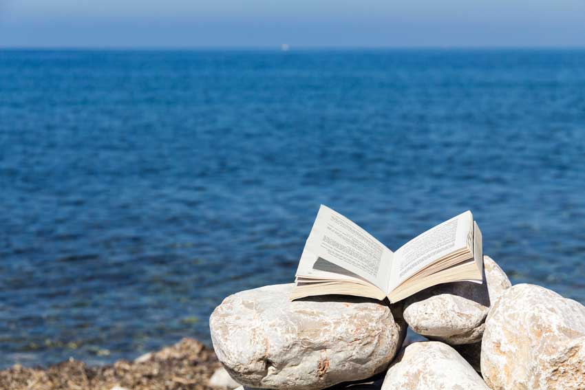 ספר פתוח על סלעים לבנים בחוף מול הים הכחול.מדגים עומק, צלילות וזרימה הרמונית בעריכה ספרותית.